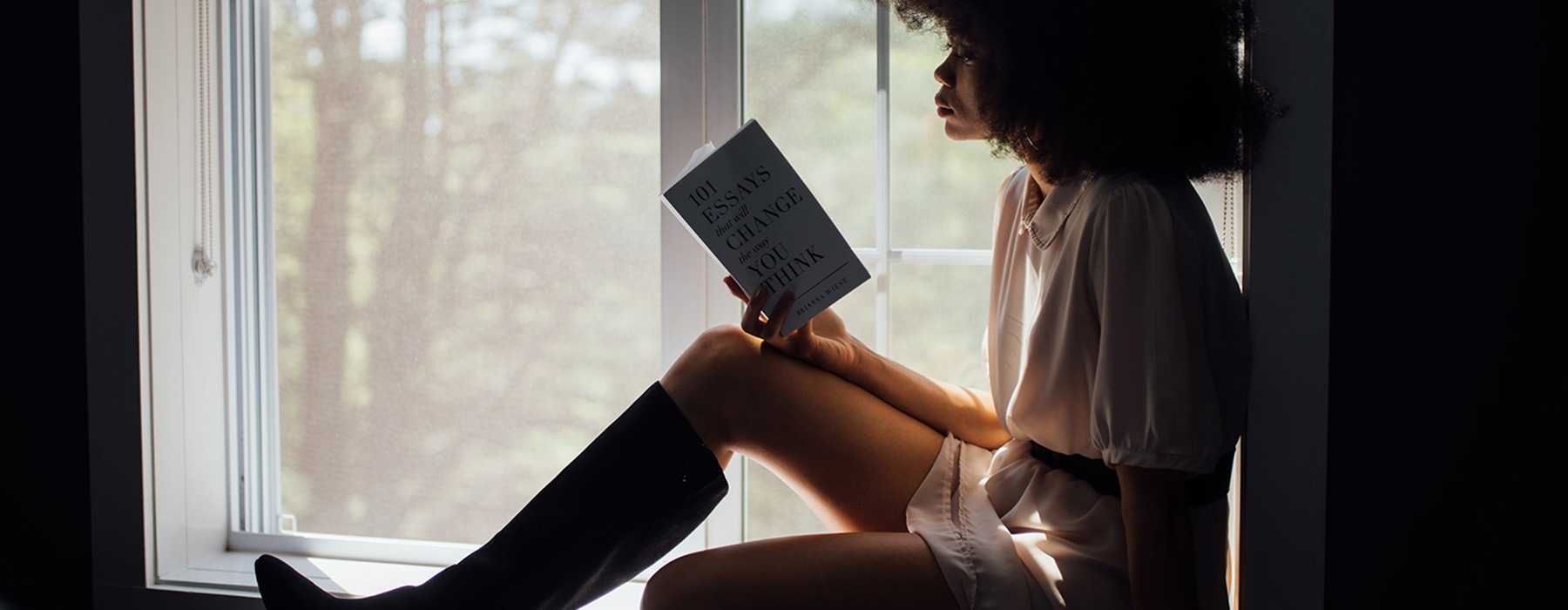 lifestyle image of a woman reading a book while sitting back in a window